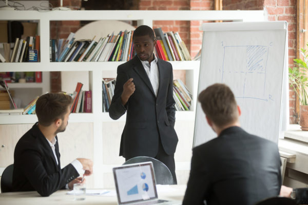 African american businessman giving presentation to partners working with flip chart at corporate meeting, black business coach explaining project strategy using whiteboard on group office training