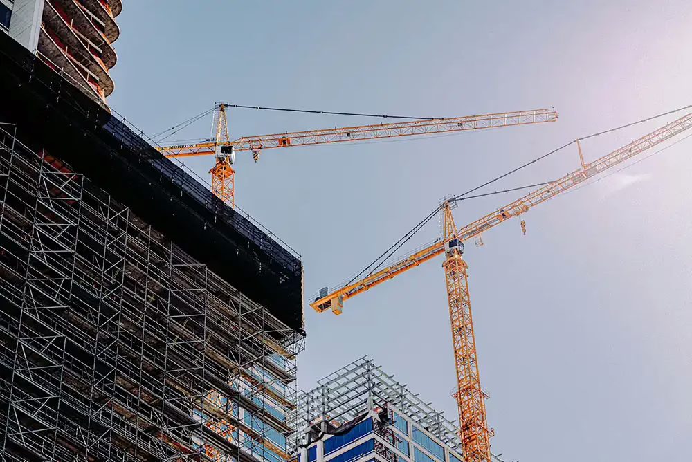 Two large construction cranes operate above high-rise buildings under construction against a clear sky